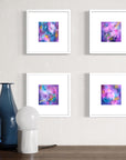 Four square abstract art prints with colorful purple, pink, and blue cosmic designs, each in a white frame with a white mat, arranged in a 2x2 grid on a textured light gray wall above a wooden surface with a blue glass vase and a white globe lamp.
