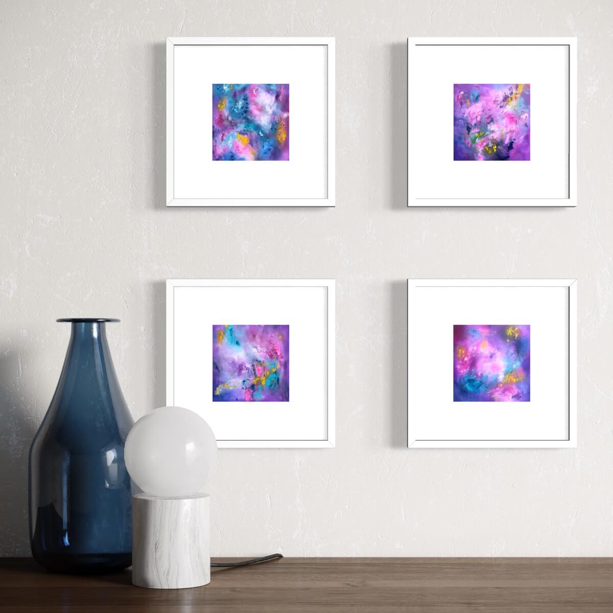 Four square abstract art prints with colorful purple, pink, and blue cosmic designs, each in a white frame with a white mat, arranged in a 2x2 grid on a textured light gray wall above a wooden surface with a blue glass vase and a white globe lamp.