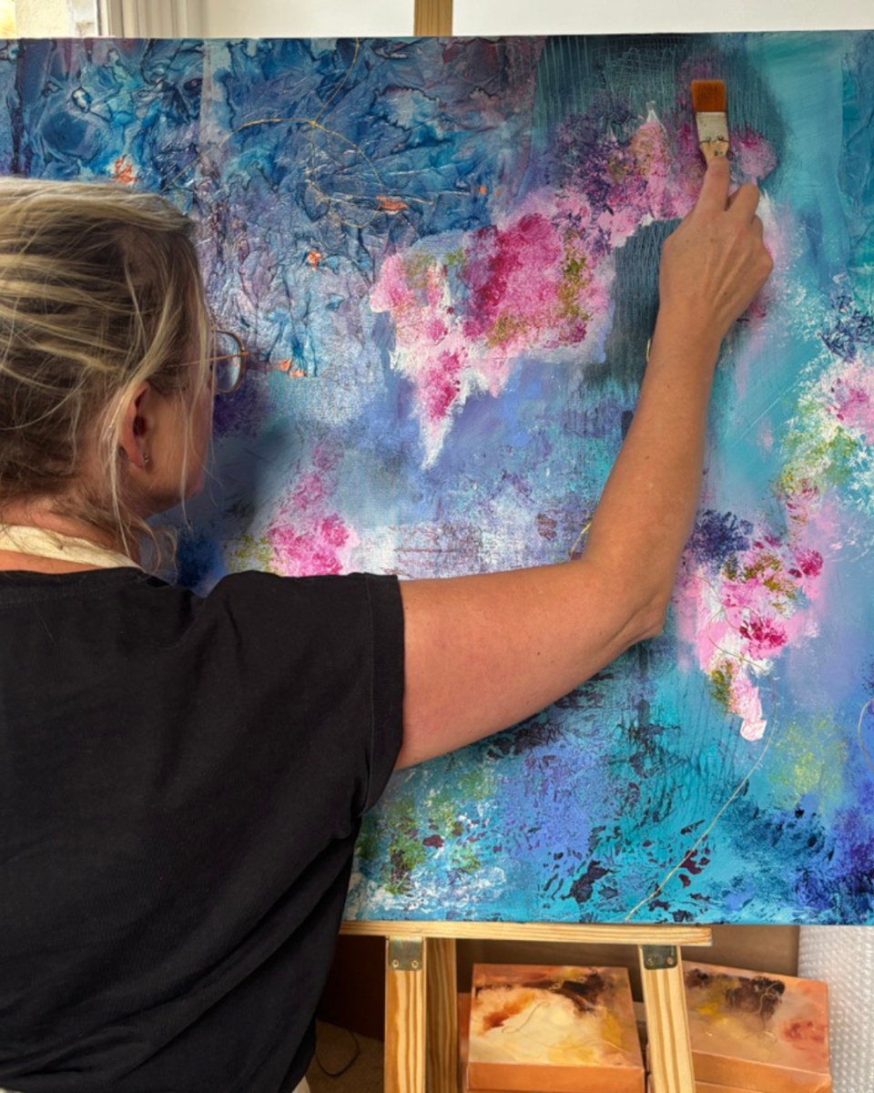 A woman painting on a large canvas with blue, pink, and teal abstract brushstrokes, standing behind an easel in an indoor studio setting.